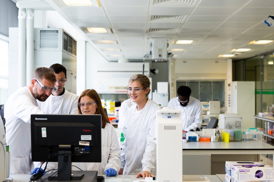 Group of people in white lab coats and wearing safety glasses, gathered round a computer