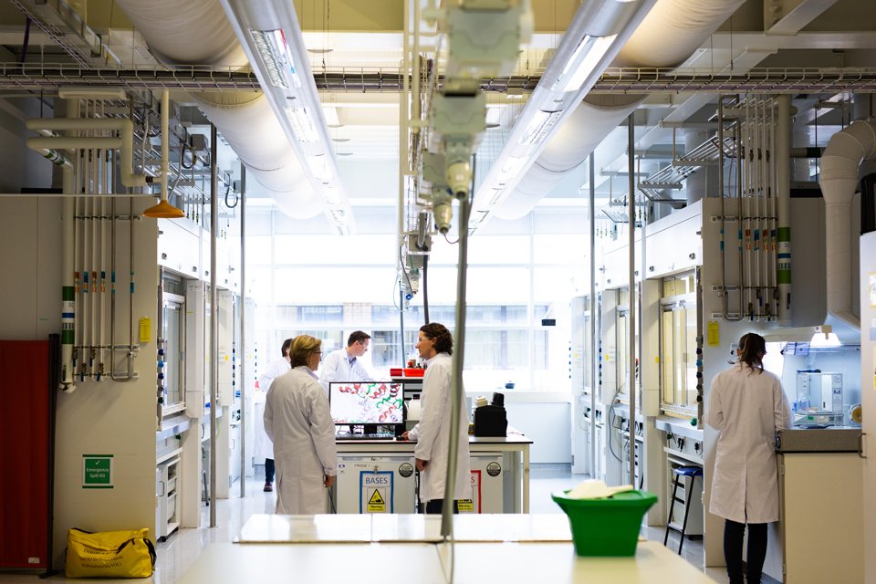 Man and woman wearing white lab coats in a lab space, collaborating by a computer in the middle of the room surrounded by lab equipment