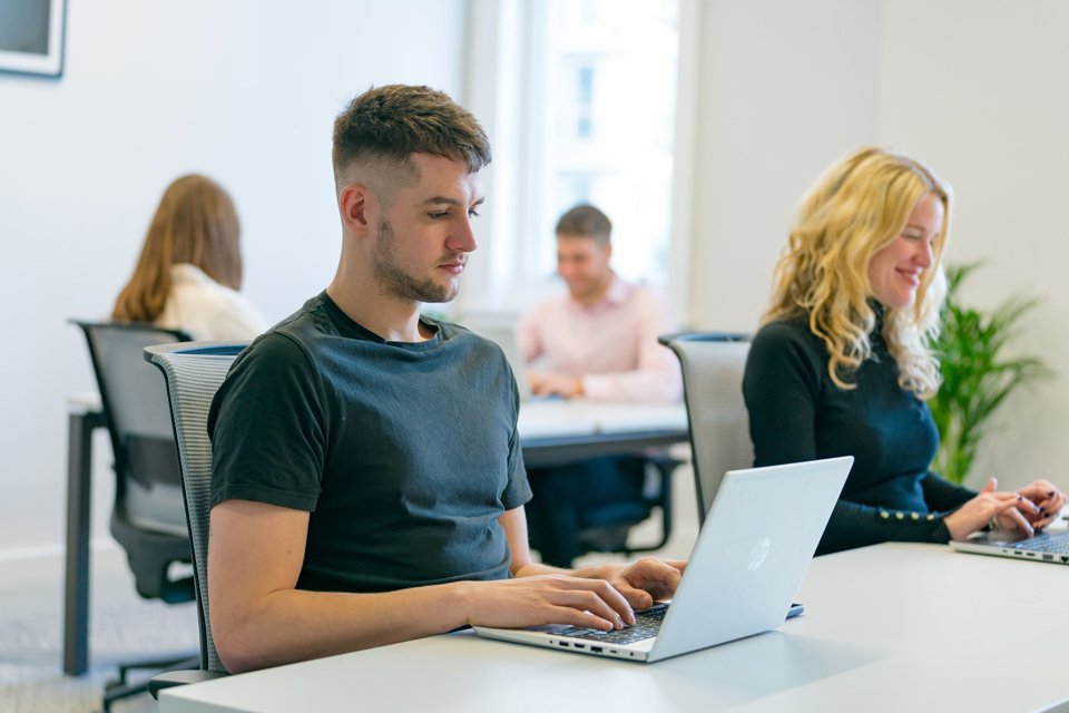 Man and a woman in the foreground sat at a white desk working on laptops.