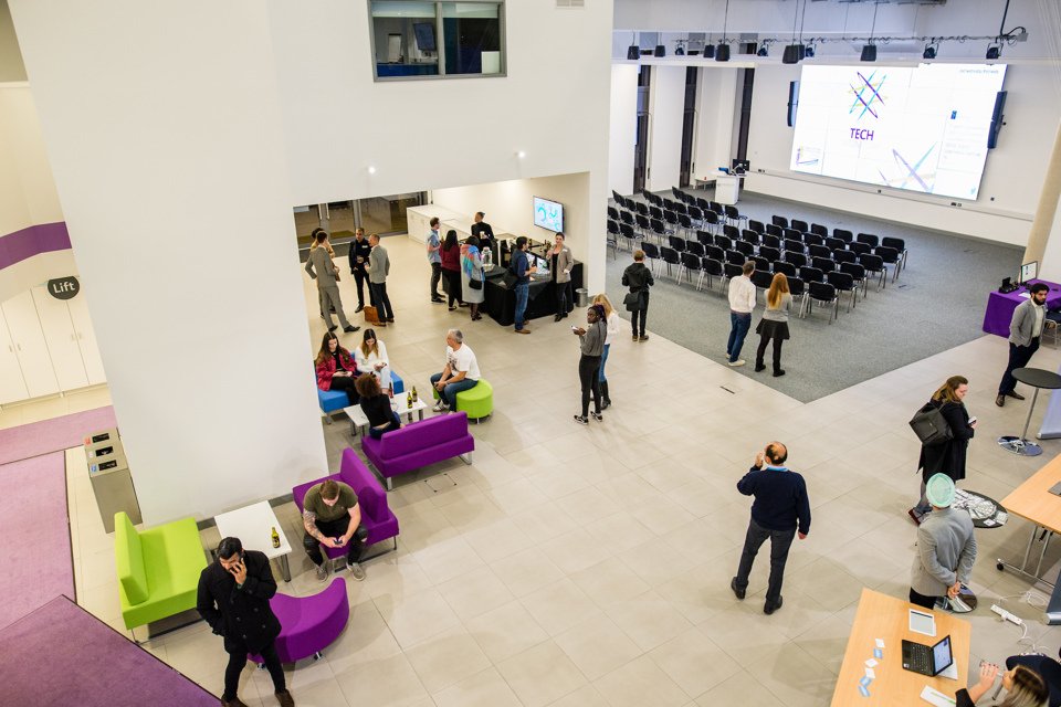 An aerial view of one of Innovation Birmingham's meeting spaces showing people stood talking, with chairs set out in a conference layout with a big screen projector.