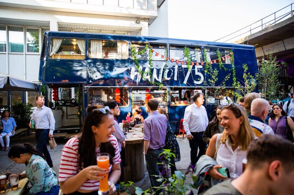 Groups of people socialising at different tables in a beer garden near a bus converted into a bar