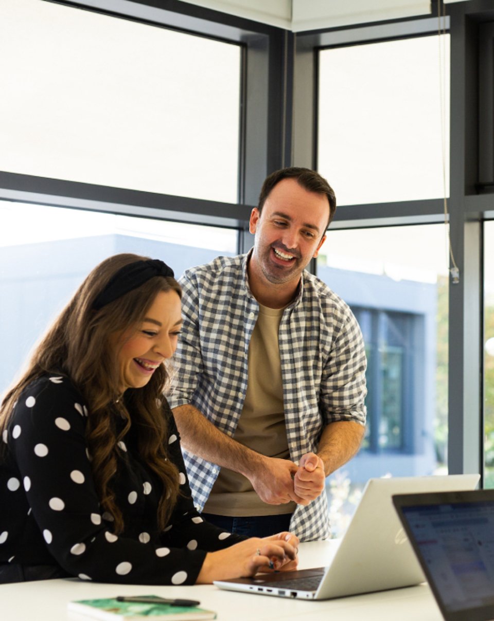 A man and a woman working on a laptop and smiling near a large window in an office