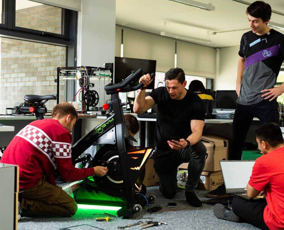 4 people working on a modified spinning bicycle with tools on the floor and looking at a laptop