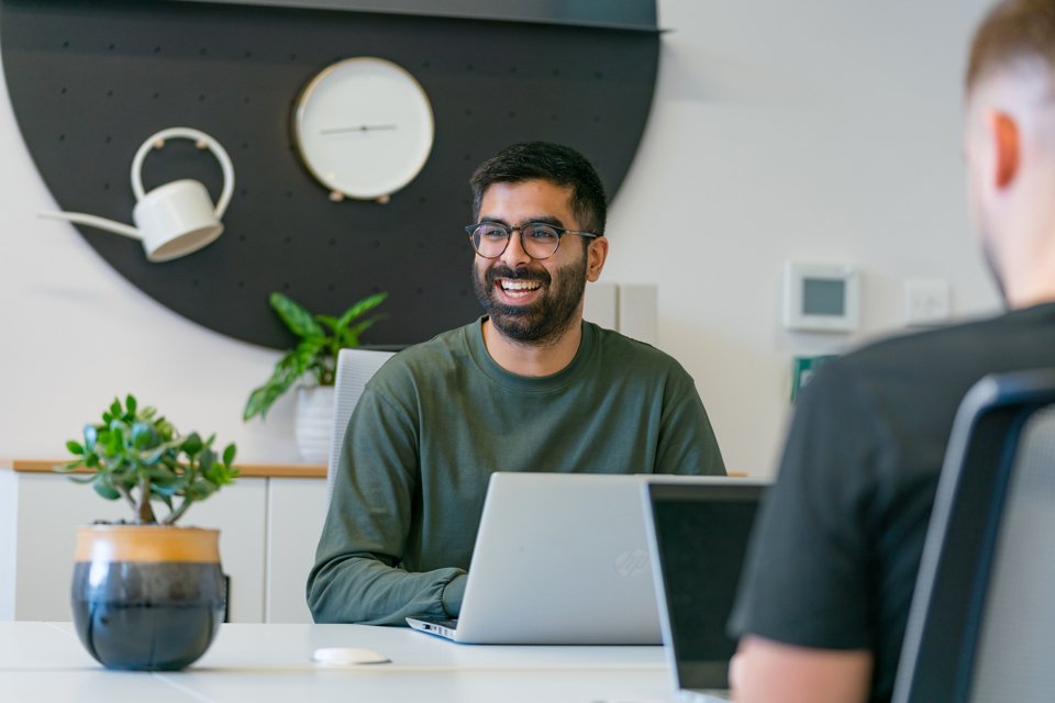 Man sat at a desk with a laptop in a white themed room and plants, looking to his right and laughing