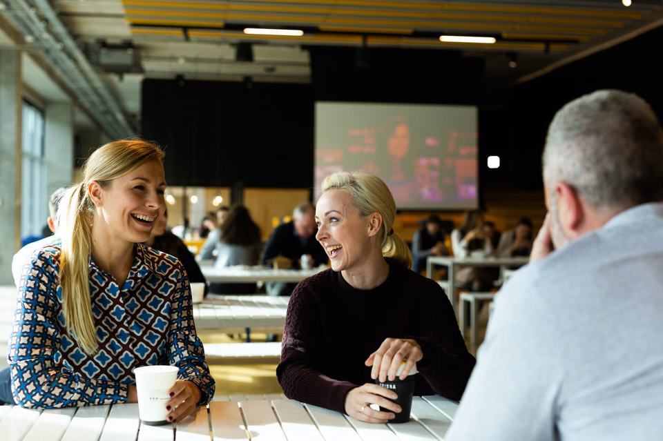 A group of 3 people sat down smiling, having a conversation while drinking a coffee