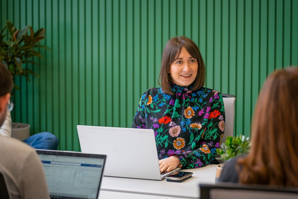 Person sat at a desk with a laptop having a discussion with another person