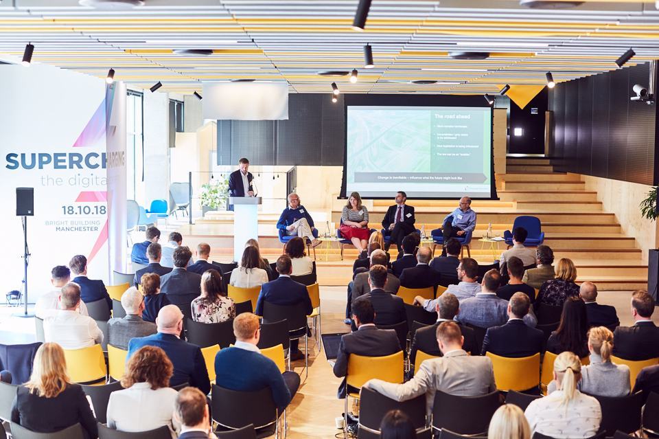 Crowd of people sat down at a conference listening to a panel of four speakers