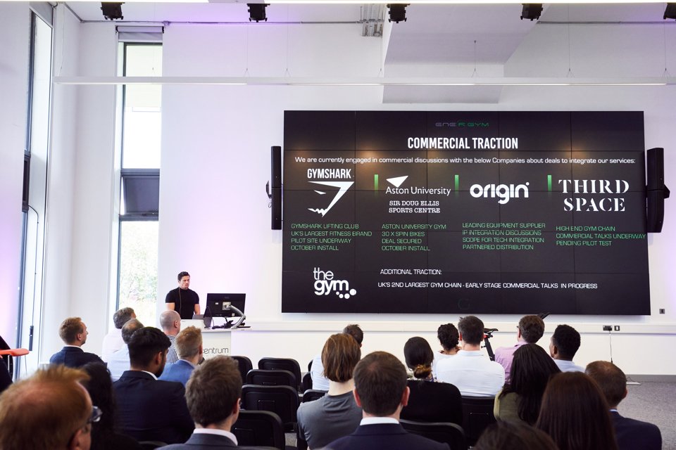 A group of people watching a large-screen presentation from Energym detailing the commercial traction they have made through networking