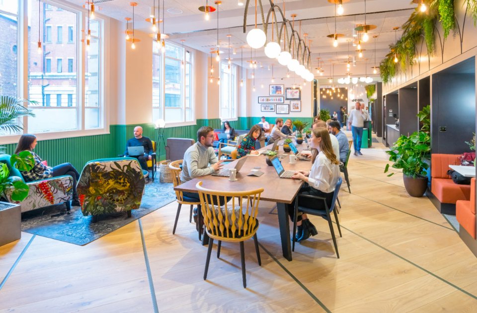 Crowd of people sat at a long desk and comfy chairs collaborating and working in a brightly lit and colourful room