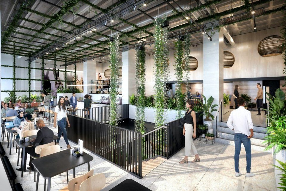 Crowd of people walking around and sat working in a large spacious area with plant decor hanging from the ceiling and a stair case in the middle of the room