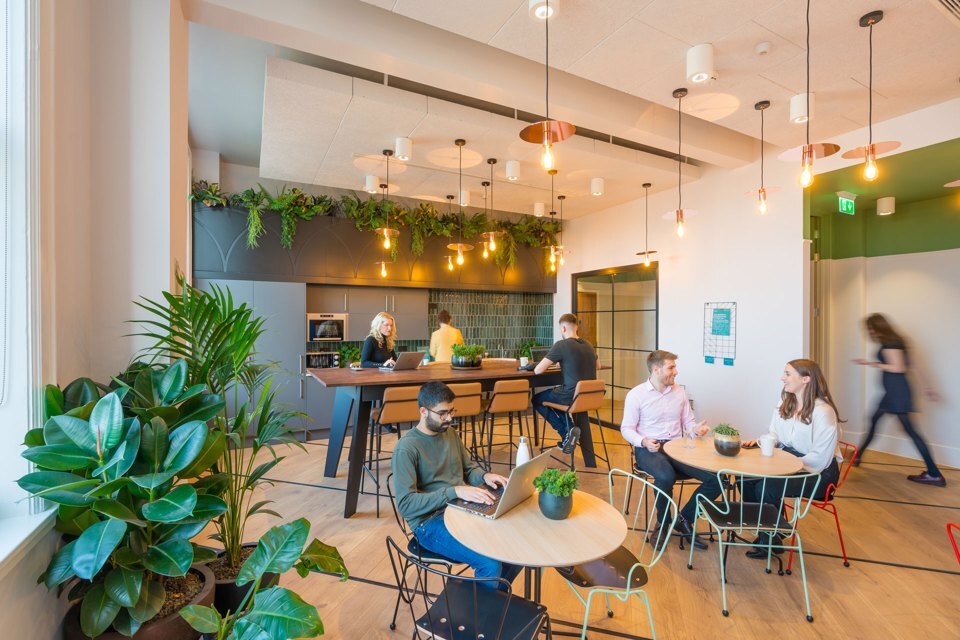 Group of people sat around a kitchen area working on laptops and chatting to one another