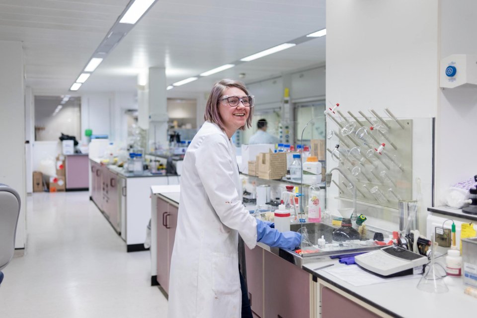 Woman in a white lab coat, wearing blue gloves and safety glasses, stood by a lab desk and smiling