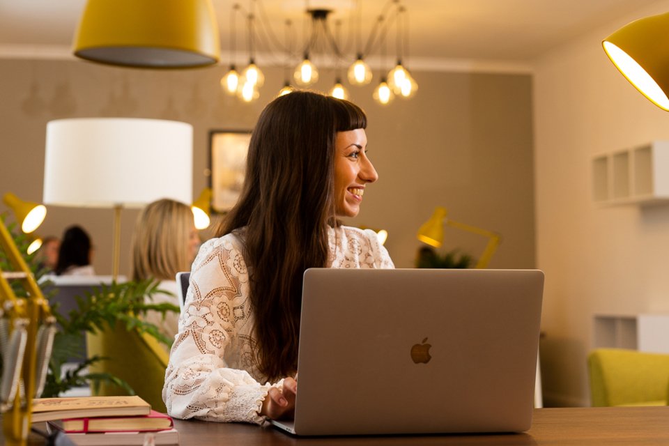 Woman sat in large chill out area, with ambient lighting, looking to her left and smiling while working on a laptop