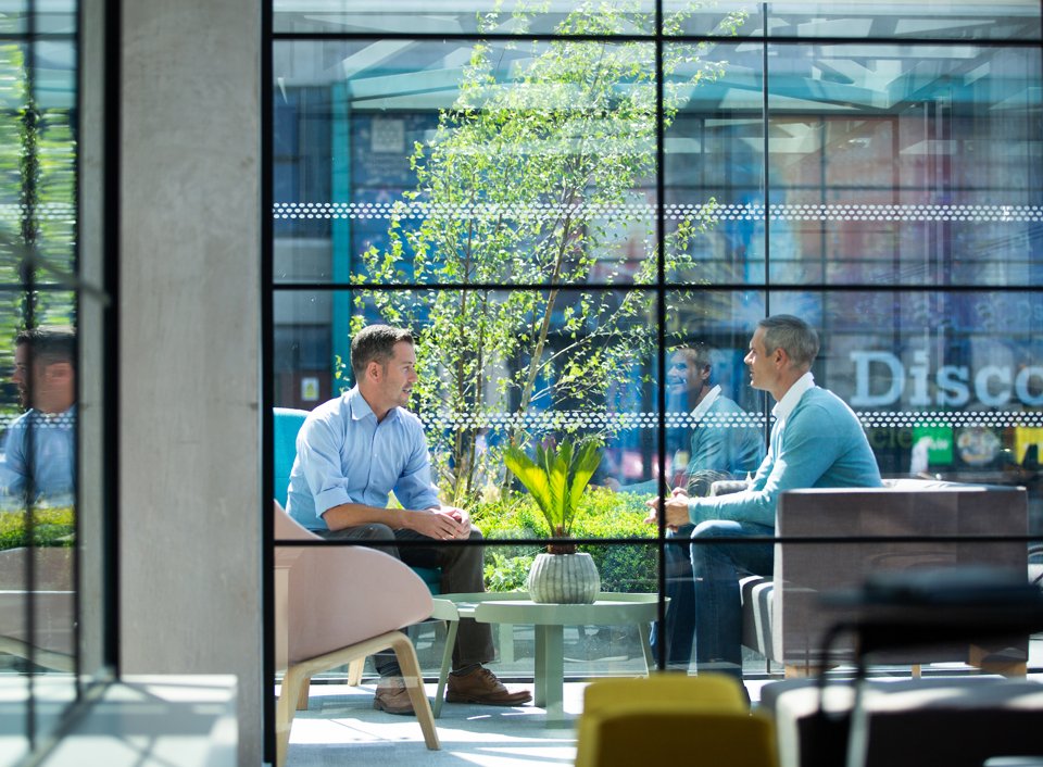 Two men in blue shirts sat around a small coffee table and talking in a large, daylight-lit meeting space