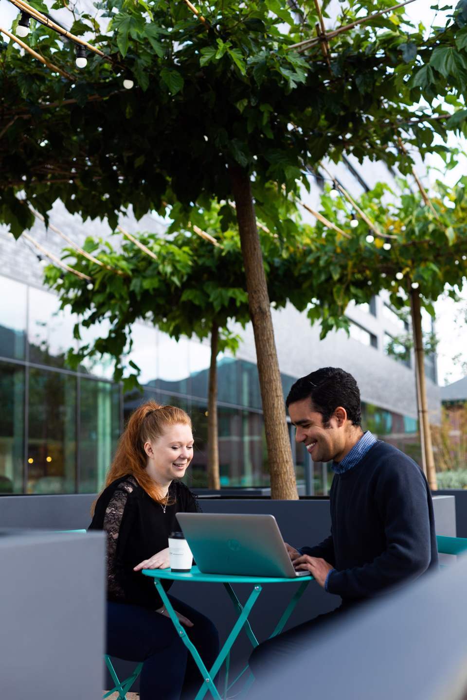 Man and a woman sat at a desk outside smiling while working on a laptop