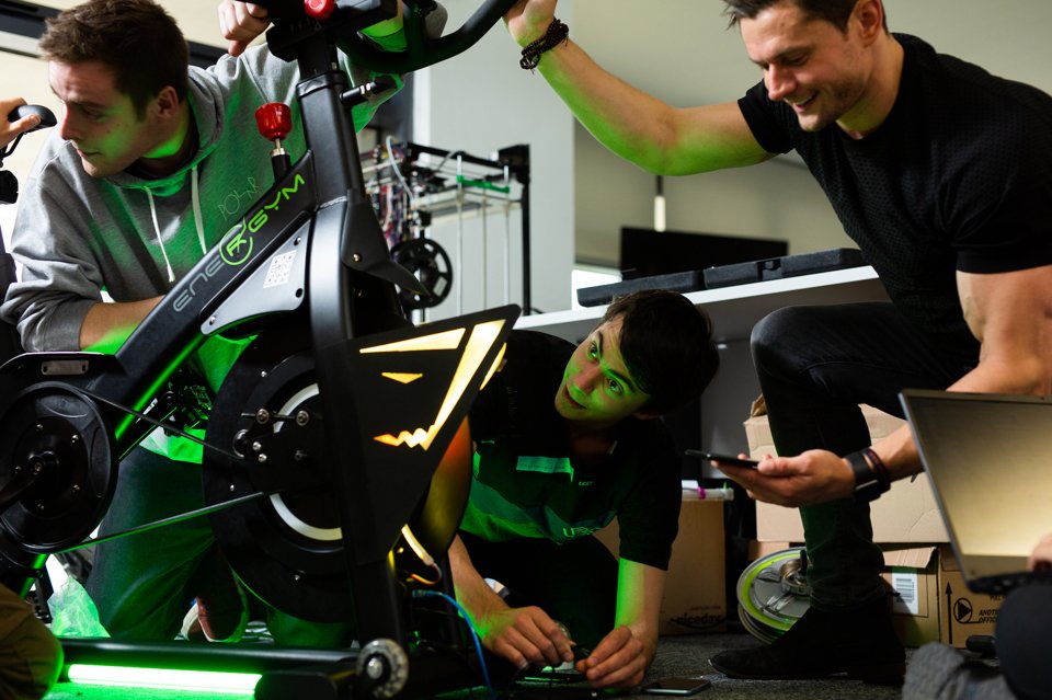 Three men gathered around a customised spinning bike whilst holding wires together and looking at data on their smartphone